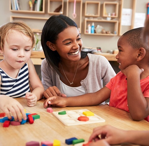 Junge Frau sitzt mit zwei kleineren Kindern an einem Tisch., sie machen ein Puzzle mit Holzsteinen.